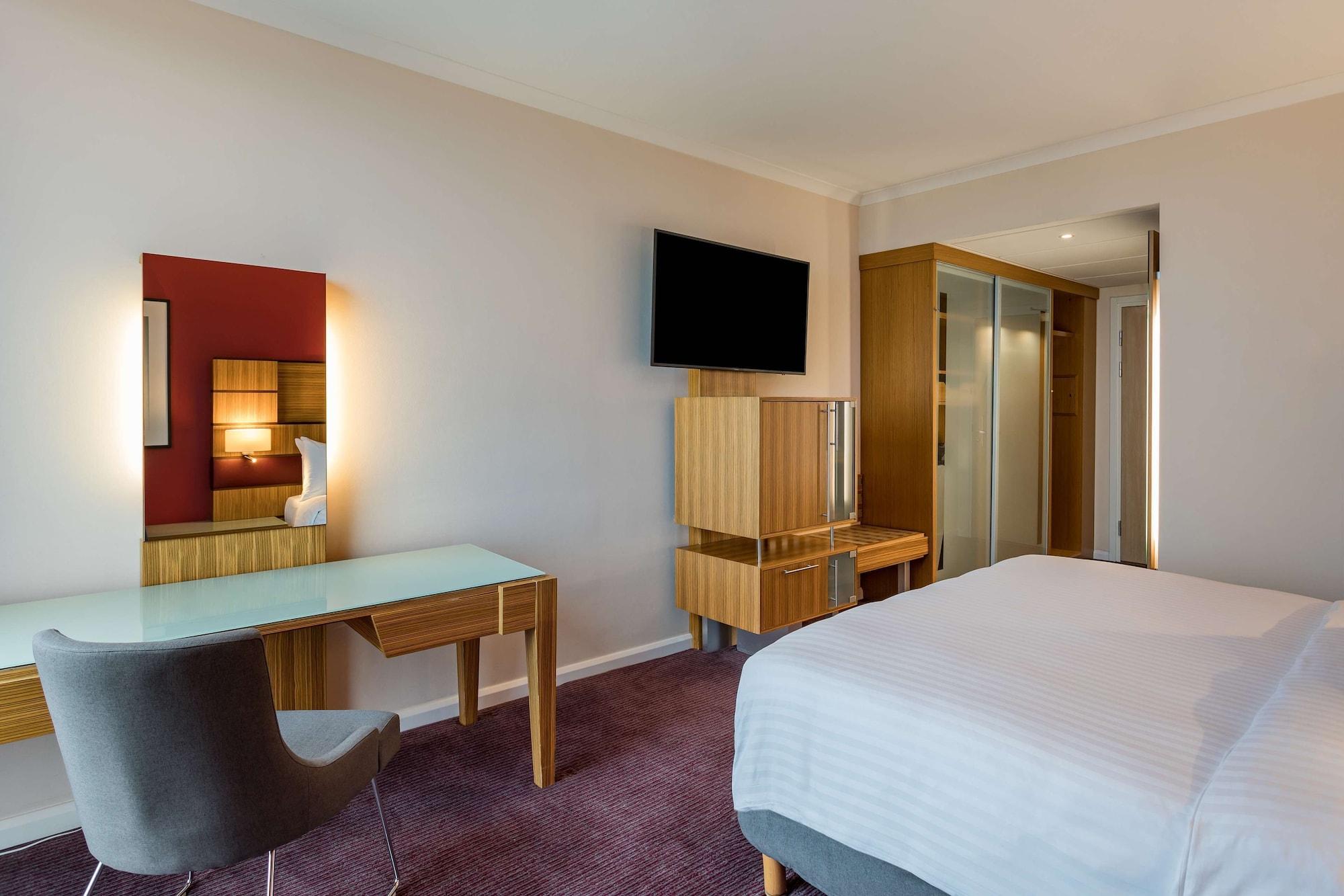 Radisson Blu Hotel London Stansted Airport Stansted Mountfitchet Exteriér fotografie The photo shows a modern hotel room. It features a bed with white bedding, a flat-screen television mounted on the wall, and a contemporary desk with a chair. There is also a mirror on the wall above the desk, and a wooden unit that appears to have s